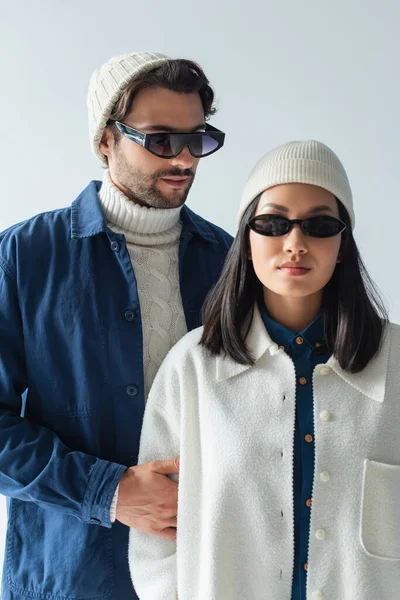 Young asian woman in white beanie and dark sunglasses near man in blue jacket isolated on grey — Stock Photo