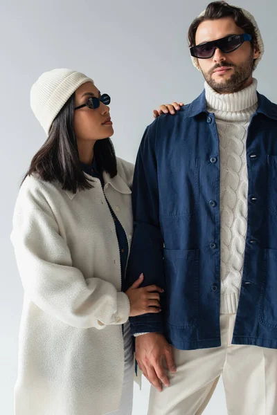 Mujer asiática en chaqueta blanca y gafas de sol oscuras mirando hombre elegante aislado en gris - foto de stock