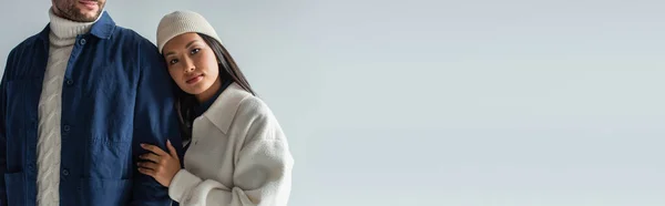 Bonita mujer asiática en chaqueta blanca y gorro apoyado en el hombre aislado en gris, bandera - foto de stock