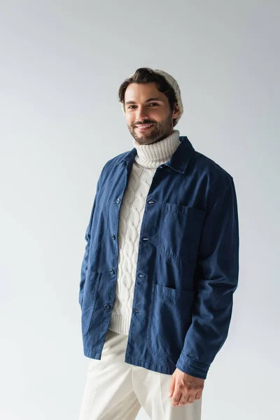 Hombre feliz en chaqueta azul y suéter de punto blanco sonriendo a la cámara aislado en gris - foto de stock