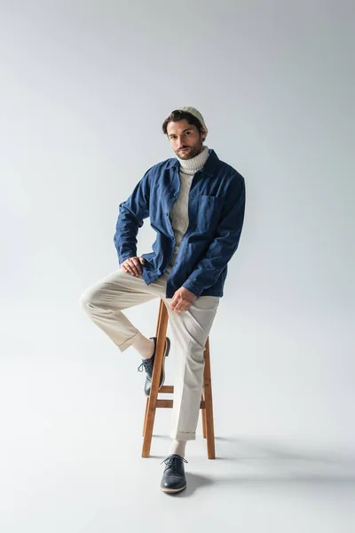 Vista completa del hombre en chaqueta azul y pantalones blancos sentados en taburete alto en gris - foto de stock