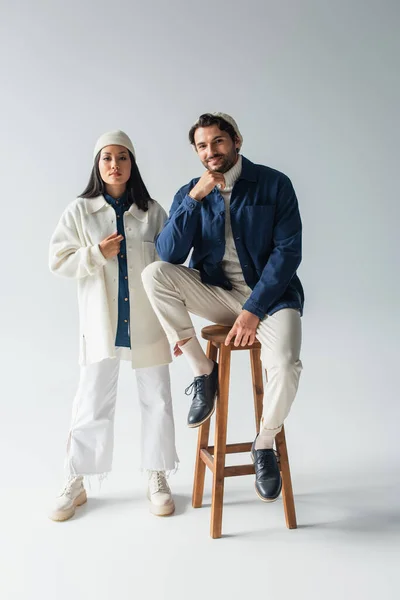 Full length view of trendy asian woman near sorrindo man sitting on high stool on grey — Fotografia de Stock