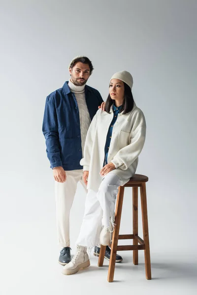 Stylish man looking at camera near asian woman sitting on high stool on grey — Stock Photo