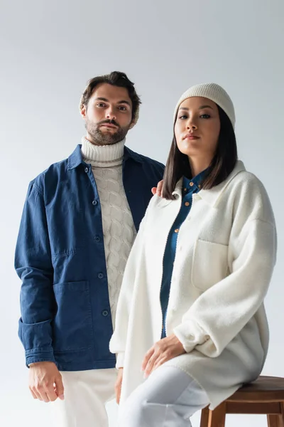 Hombre de moda mirando a la cámara cerca de mujer asiática en chaqueta blanca y gorro aislado en gris - foto de stock