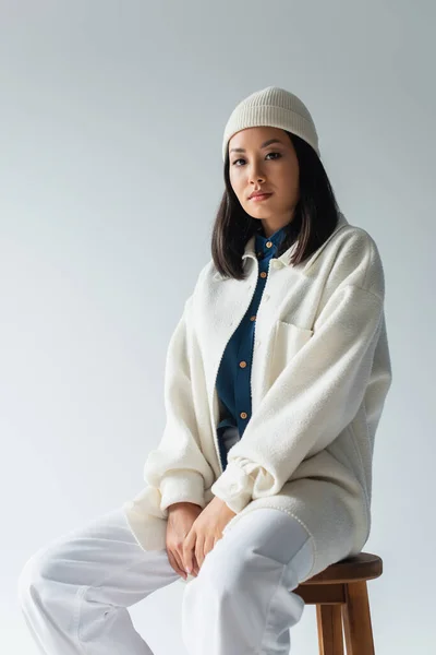 Hübsche asiatische Frau in weißer Mütze und Jacke sitzt auf einem Hocker und schaut vereinzelt in die Kamera auf grau — Stockfoto