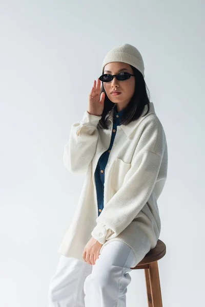 Молода азіатка в білому піджаку і бані, торкаючись темних сонцезахисних окулярів, ізольована на сірому — Stock Photo