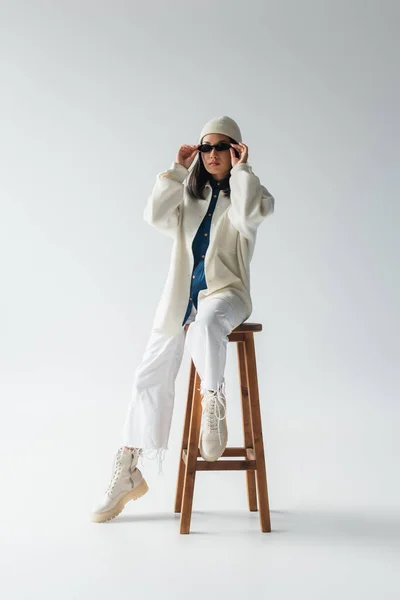 Full length view of asian woman in white clothes and black sunglasses posing on high stool on grey — Fotografia de Stock