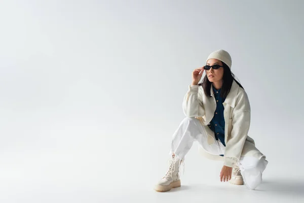 Vista completa de mujer asiática en ropa blanca ajustando gafas de sol y mirando hacia otro lado en gris - foto de stock