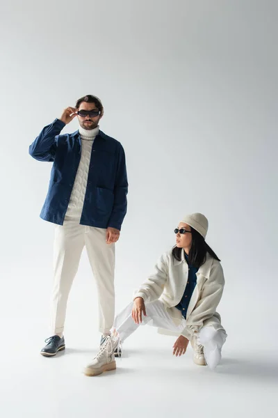 Vista completa de hombre de moda ajustando gafas de sol oscuras cerca de mujer asiática en gris - foto de stock