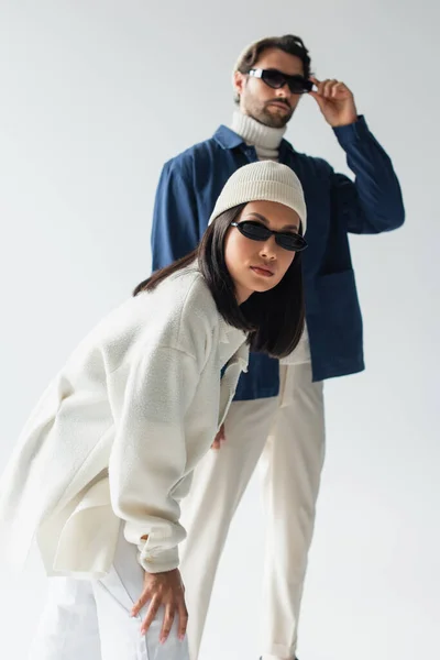 Asian woman in white jacket and dark sunglasses looking at camera near blurred man on grey — Stock Photo