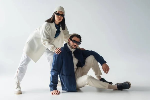 Pareja interracial de moda en gafas de sol oscuras mirando a la cámara mientras posan en gris - foto de stock