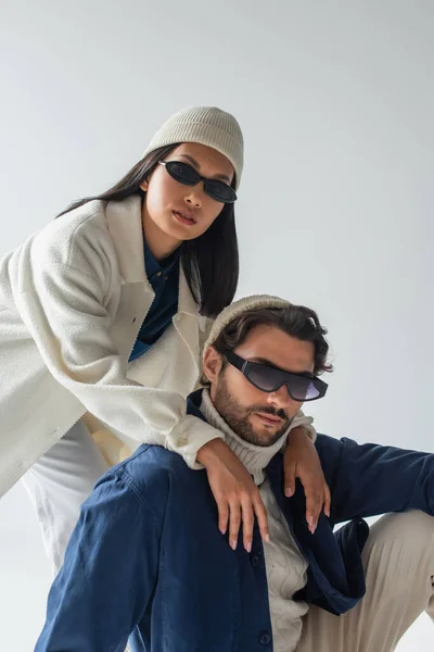 Pareja interracial de moda en gafas de sol oscuras y frijoles mirando a la cámara aislada en gris - foto de stock