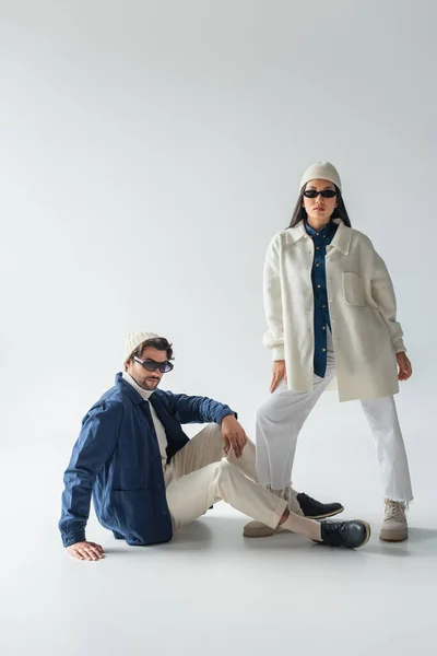 Asiatique femme en blanc vêtements et sombre lunettes de soleil posant près de l homme assis sur gris — Photo de stock