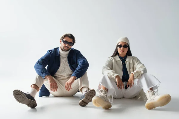 Vista completa de pareja multiétnica en ropa de moda y gafas de sol oscuras sentadas en gris - foto de stock