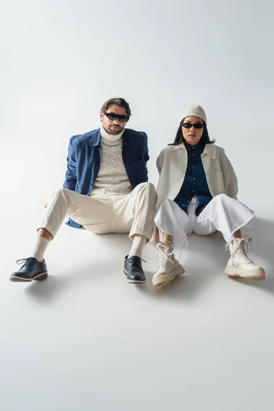 Vista a tutta lunghezza della coppia interrazziale alla moda in occhiali da sole scuri seduti sul grigio — Foto stock