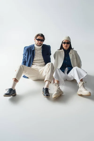 Vue pleine longueur du couple interracial à la mode dans des lunettes de soleil sombres assis sur le gris — Photo de stock