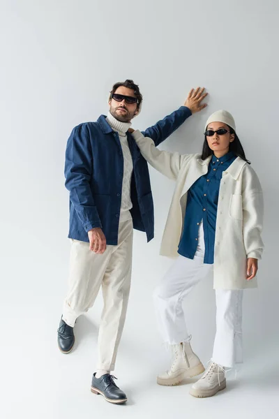 Vue pleine longueur de couple interracial élégant en blanc et bleu vêtements et lunettes de soleil sur gris — Photo de stock