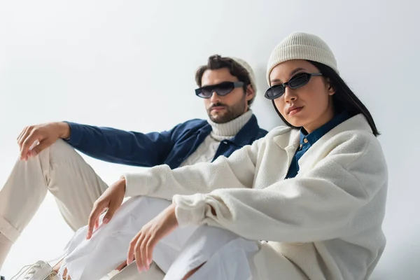 Модная азиатская женщина в темных солнцезащитных очках сидит рядом с размытым мужчиной на сером — стоковое фото