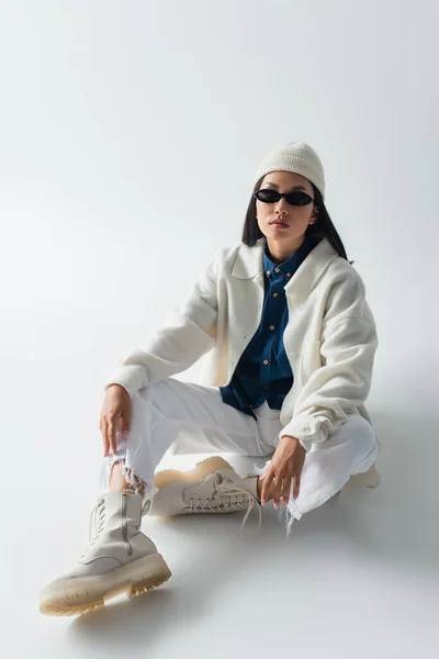 Asiatique femme en blanc vêtements et sombre lunettes de soleil regarder caméra tandis que assis sur gris — Photo de stock