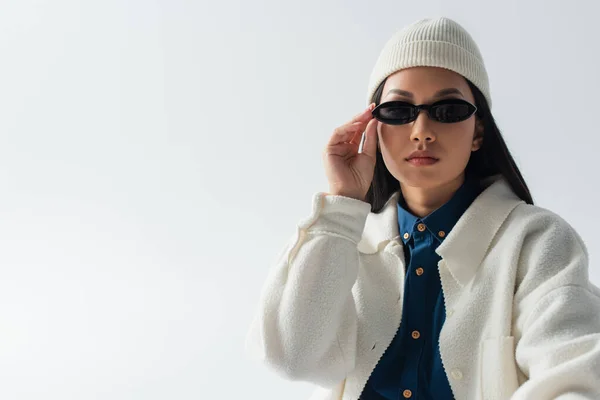 Jovem asiático mulher no branco jaqueta e gorro ajustando escuro óculos de sol isolado no cinza — Fotografia de Stock