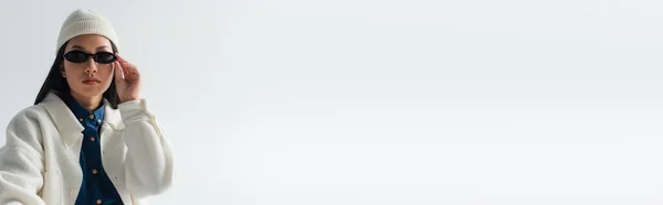 Donna asiatica alla moda in giacca bianca e berretto regolazione occhiali da sole isolati su grigio, banner — Foto stock