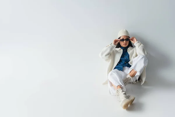 Vue grand angle de femme asiatique à la mode ajustant les lunettes de soleil tout en étant assis sur gris — Photo de stock