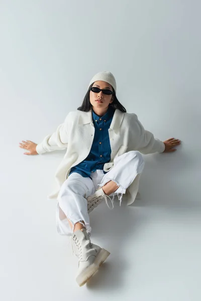 Высокий угол обзора азиатской женщины в белой одежде и темных солнцезащитных очках, сидящих на сером — стоковое фото