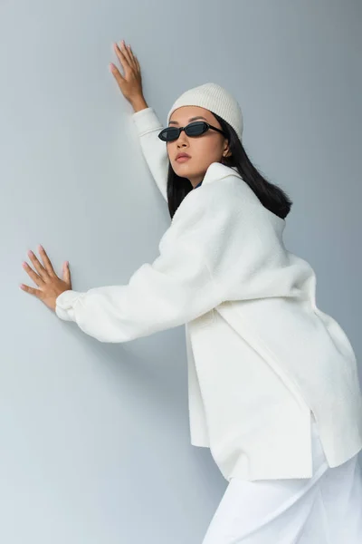Стильна азіатська жінка в білому піджаку і чорних сонцезахисних окулярах позує на сірому — стокове фото