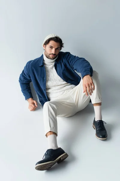 Высокий угол обзора человека в белых брюках и синей куртке, смотрящего в камеру, сидя на сером — стоковое фото