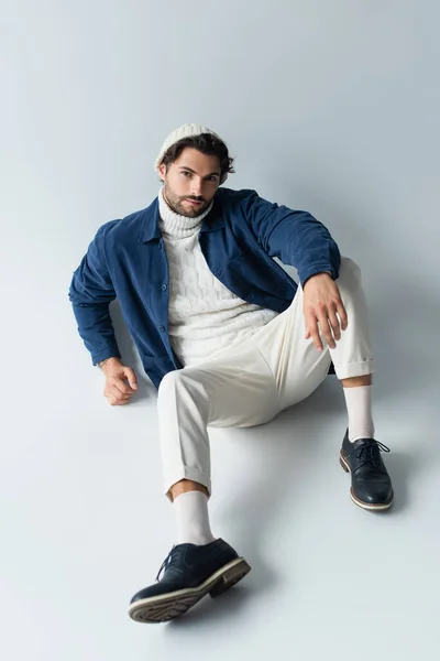 Vista de ángulo alto del hombre en pantalones blancos y chaqueta azul mirando a la cámara mientras está sentado en gris - foto de stock