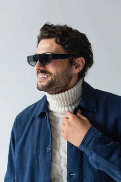 Homme heureux en veste bleue, pull blanc et lunettes de soleil sombres regardant loin isolé sur gris — Photo de stock