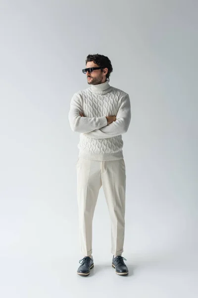 Morena hombre en pantalones blancos, suéter de punto y gafas de sol oscuras de pie con los brazos cruzados en gris - foto de stock