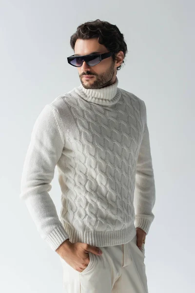 Élégant homme en pull tricoté blanc et lunettes de soleil sombres debout avec les mains dans des poches isolées sur gris — Photo de stock