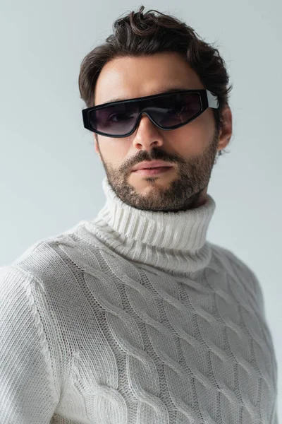 Модный мужчина в белом свитере и темных солнцезащитных очках, смотрящий на камеру, изолированную на сером — стоковое фото