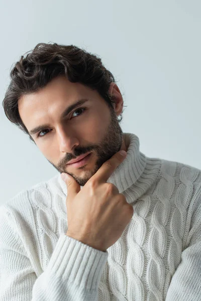 Brunette homme en chandail tricoté blanc touchant menton et regardant la caméra isolée sur gris — Photo de stock