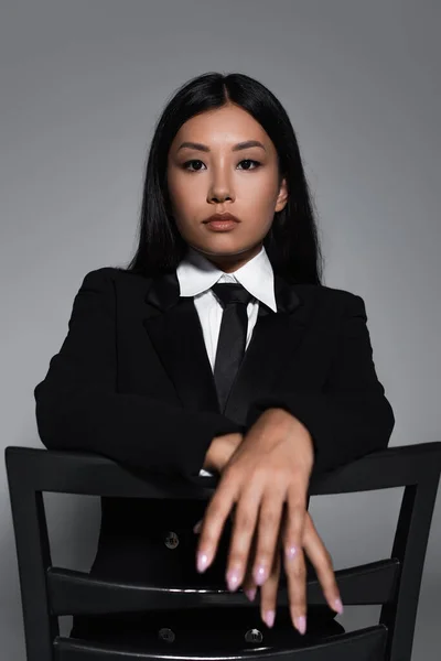 Молодая азиатская женщина в черной формальной одежде смотрит в камеру, позируя на стуле, изолированном на сером — стоковое фото
