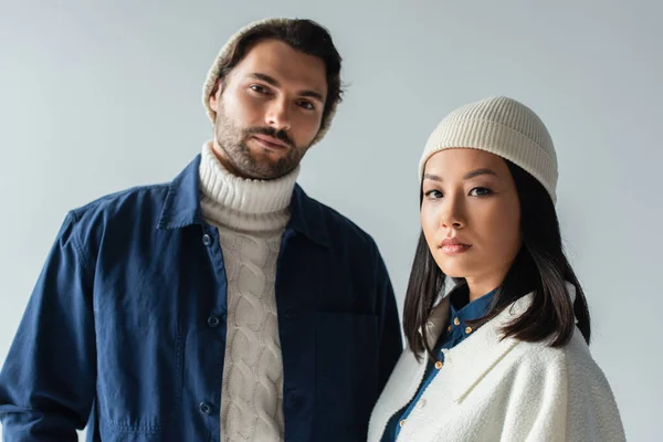 Casal multiétnico em roupas elegantes e beanies olhando para a câmera isolada em cinza — Fotografia de Stock