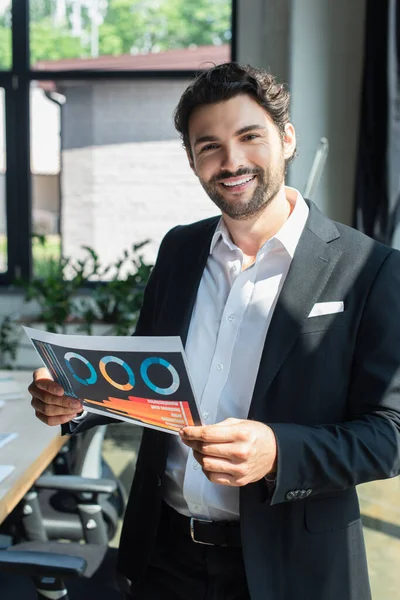 Elegante hombre de negocios en blazer negro y camisa blanca con infografías en la oficina - foto de stock