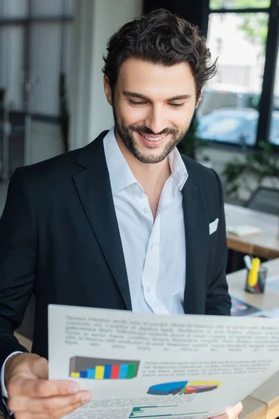 Uomo d'affari sorridente in camicia bianca e blazer nero guardando analytics in ufficio — Foto stock