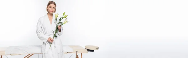 Jeune femme blonde tenant des fleurs près de la table de massage sur blanc, bannière — Photo de stock