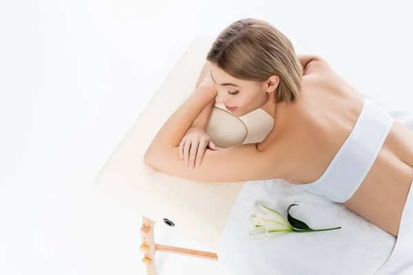 Vista ad alto angolo di felice giovane donna con spalle nude sdraiato sul tavolo da massaggio vicino al fiore isolato su bianco — Foto stock