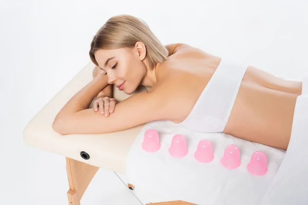 Vista de ángulo alto de la mujer joven y complacida tumbada en la mesa de masaje cerca de tazas de masaje rosa aislado en blanco - foto de stock