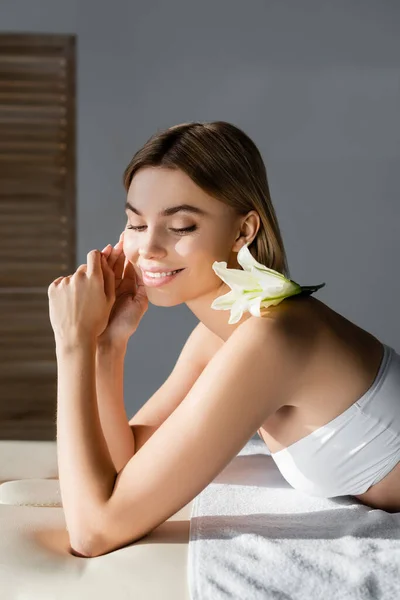 Солнце на лице довольной молодой женщины с цветком на обнаженном плече, лежащем на массажном столе — стоковое фото