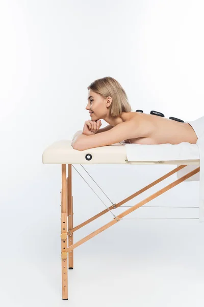 Vue latérale de heureuse jeune femme recevant un massage aux pierres chaudes sur blanc — Photo de stock