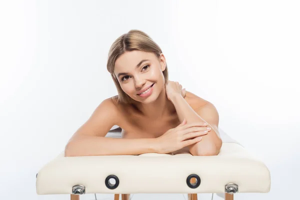 Mujer rubia positiva con los hombros desnudos acostado en la mesa de masaje en blanco - foto de stock