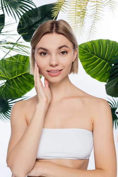 Pleased young woman with bare shoulders near green tropical leaves on white — Stock Photo