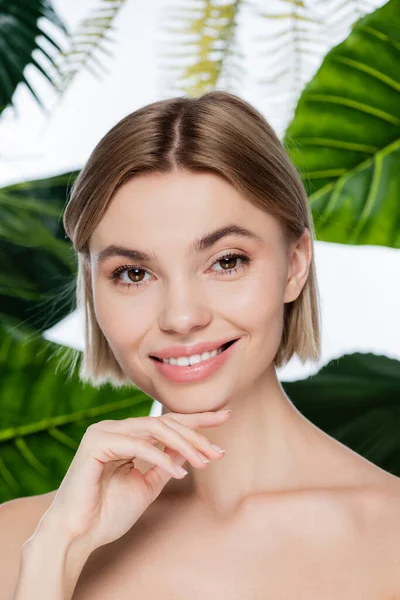 Freudige junge Frau mit perfekter Haut in der Nähe grüner tropischer Blätter auf weißem Grund — Stockfoto