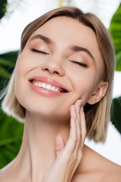 Jovem alegre tocando pele perfeita perto de folhas tropicais verdes no branco — Fotografia de Stock