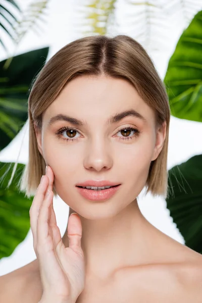 Giovane donna con pelle perfetta guardando macchina fotografica vicino foglie di palma verde su bianco — Foto stock