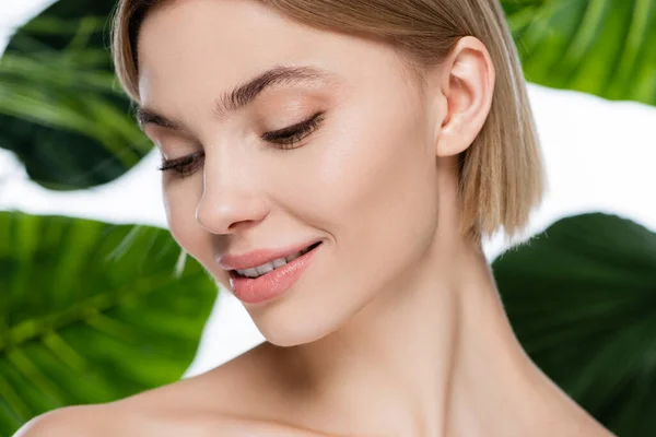 Primer plano de mujer joven complacida con la piel perfecta cerca de hojas de palma verde sobre blanco - foto de stock